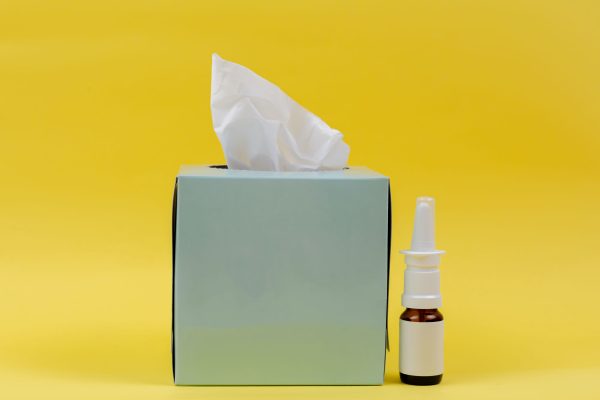 Foto van een tissuedoos met een flesje neusspray tegen een gele achtergrond