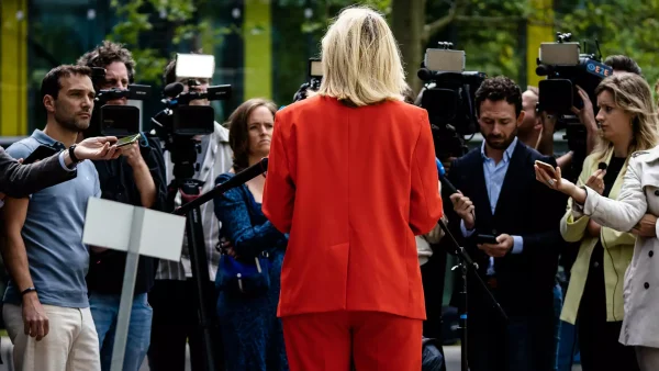 Sigrid Kaag staat met haar rug naar je toe en draagt een rood pak terwijl ze een groep journalisten te woord staat