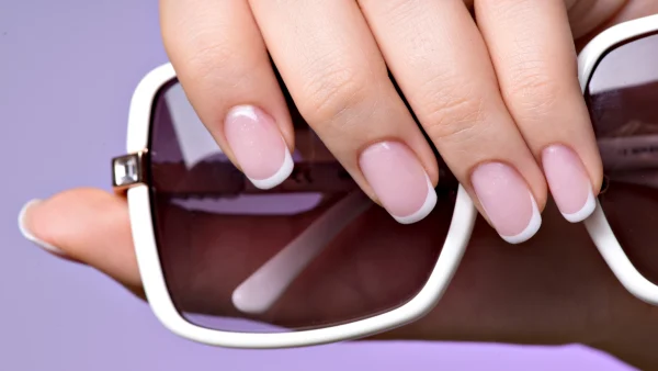 Hand met nagellak die een zonnebril vasthoudt voor een paarse achtergrond.