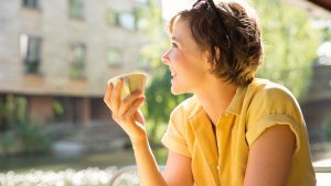 Vrouw in gele blouse zit op het terras met een kop koffie in haar hand en lacht