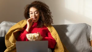 Vrouw in een rode trui kijkt kerstfilms op de bank met popcorn en een gele deken