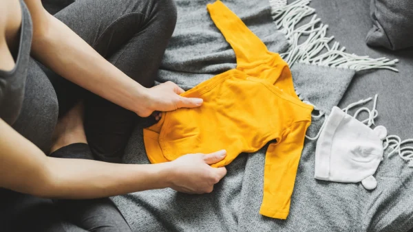 Zwangere vrouw houdt gele babykleding vast op een grijs bed