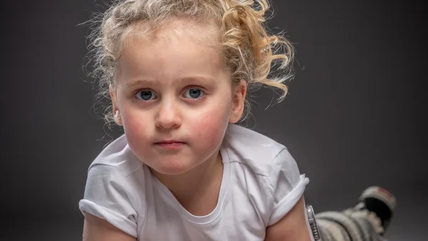 Nora, een meisje van 3 met blonde krullen. ligt op haar buik en kijkt met een neutrale blik in de camera