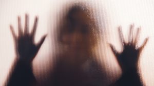 Een vrouw staat met twee handen tegen ht glas van een voordeur, gezien vanaf de buitenkant | Illustratie bij femicide