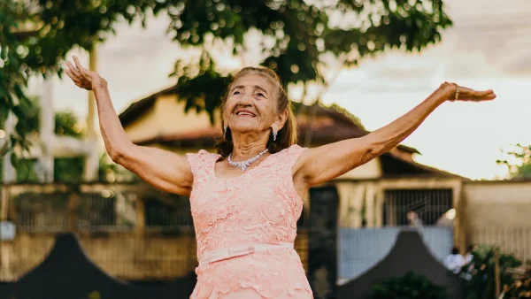 Oudere vrouw met een roze jurk heft haar handen op richting de lucht