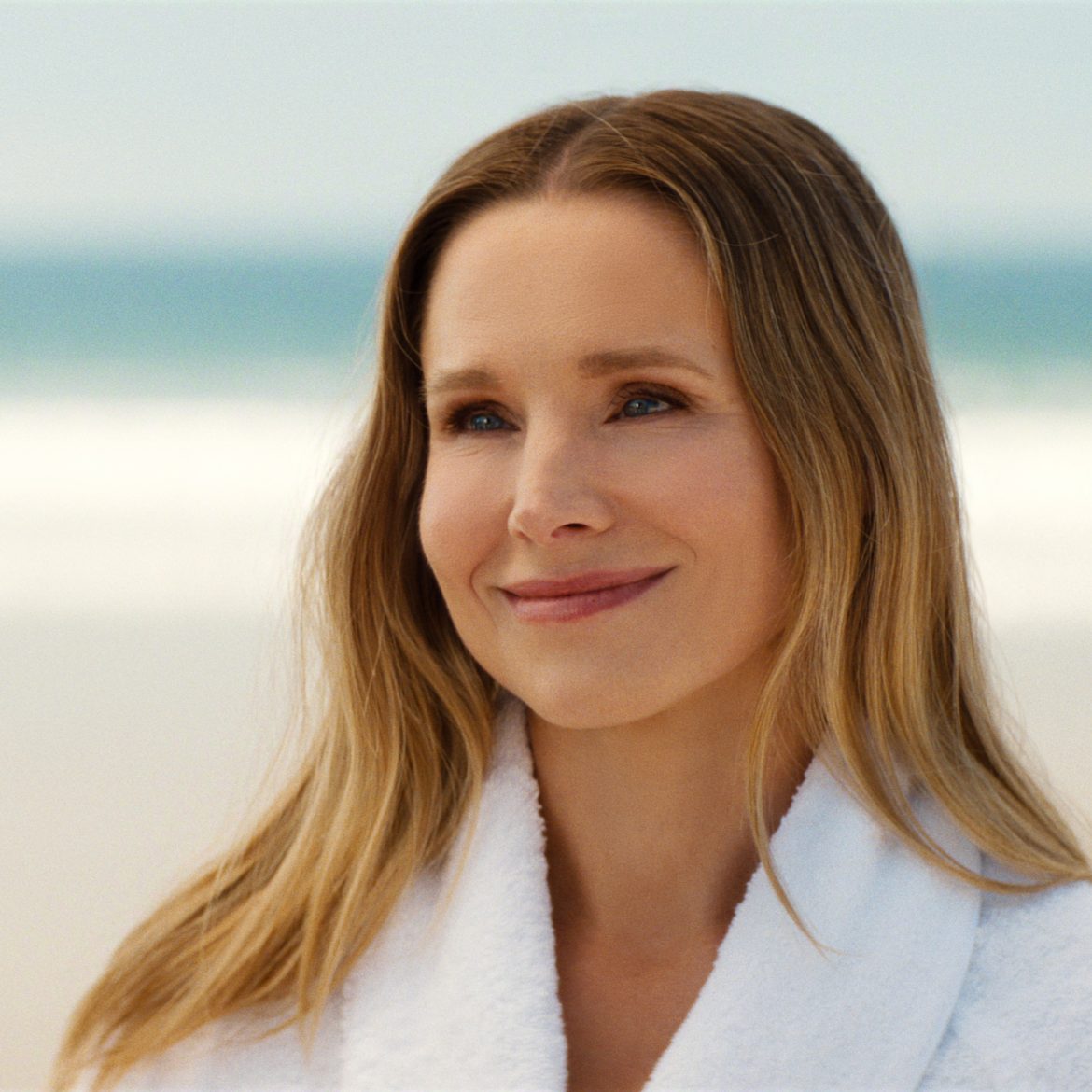 "A woman in a white robe smiles warmly at a man on a sandy beach with ocean waves in the background, under a clear sky."