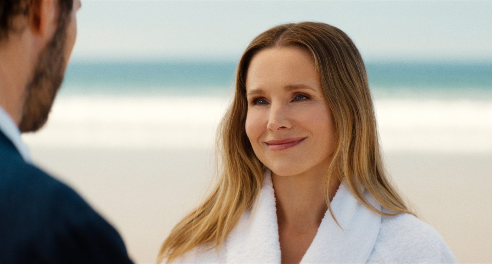 "A woman in a white robe smiles warmly at a man on a sandy beach with ocean waves in the background, under a clear sky."